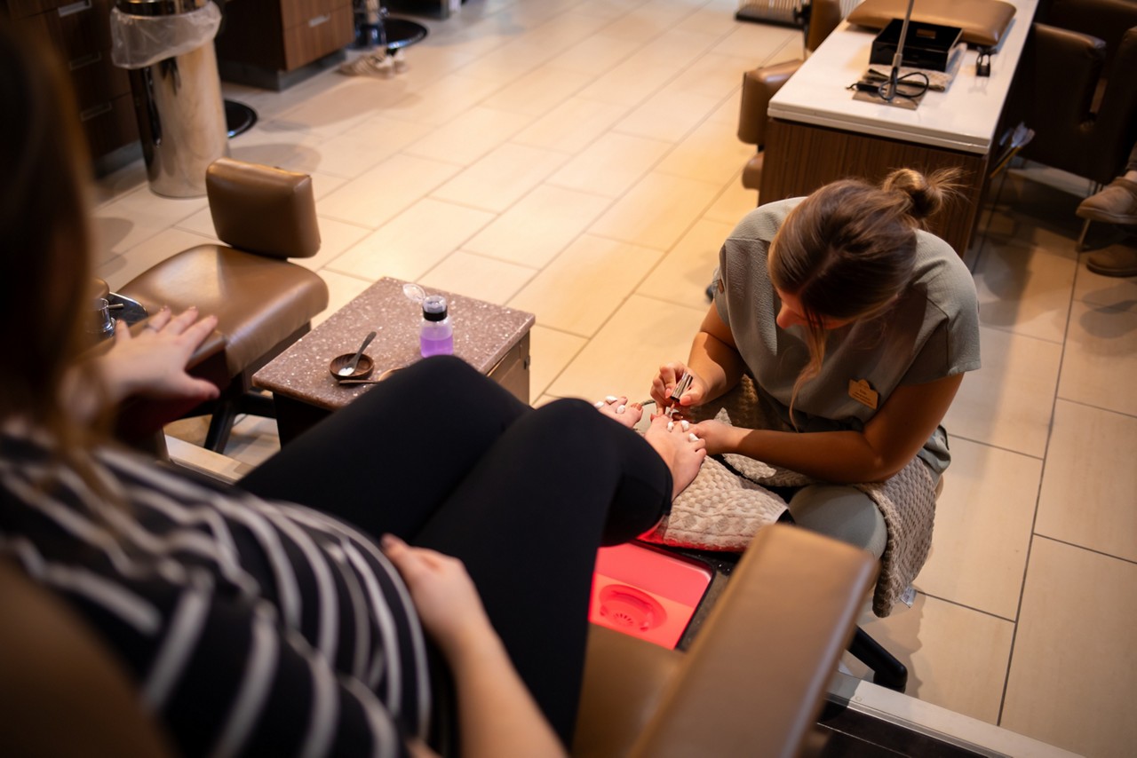 Nail technician paints client's toenails beside a pedicure chair. Client reclines with feet above glowing foot bath; small table holds polish remover, tools, and towels in a salon.
