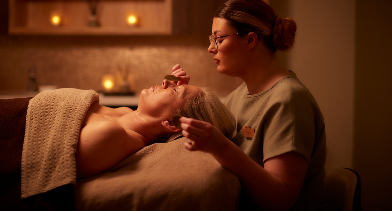 Therapist massages a client's face with smooth stones, gliding over cheeks and forehead; client lies under a towel on a treatment bed in a softly lit spa room with candles.