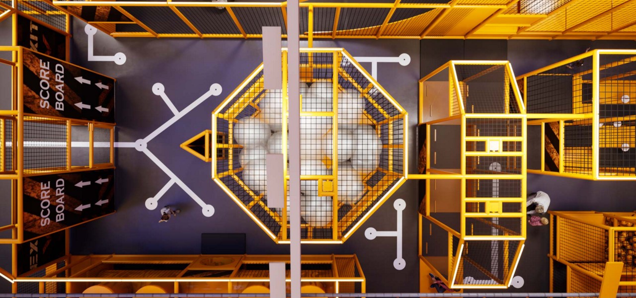 Octagonal netted pit holds white foam cubes; yellow-framed play cages surround it, overhead view with directional floor markings and signs reading SCORE BOARD, SCORE BOARD; a few people walk nearby.