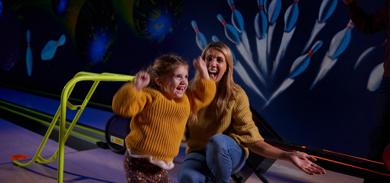 Child jumps with raised arms, celebrating. Woman kneels beside, smiling. Context: dimly lit bowling alley with neon lanes, pins mural, and a bright green bowling ramp for assistance.