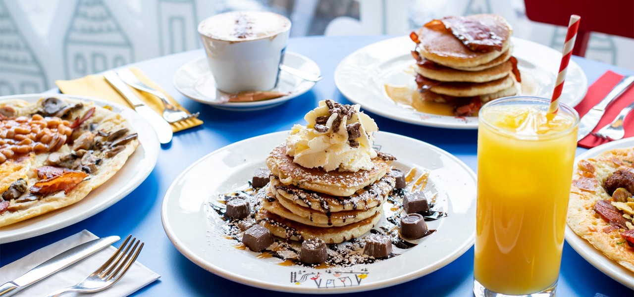 Stack of pancakes drizzled with chocolate and topped with whipped cream sits on a plate, surrounded by chocolates; nearby orange juice, coffee, and other breakfast dishes on a café table.