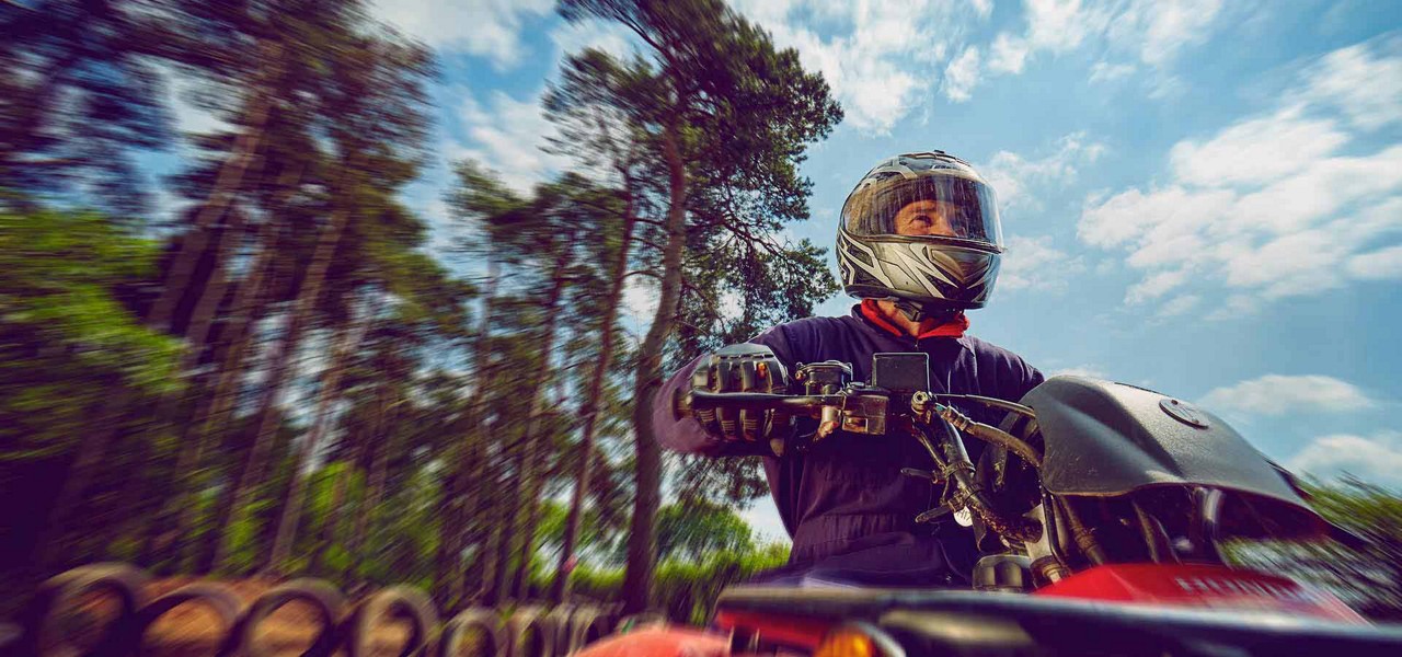 Helmeted rider controls a red ATV, accelerating along a wooded trail; motion blur streaks tall pines and blue sky, with stacked tires edging the path.