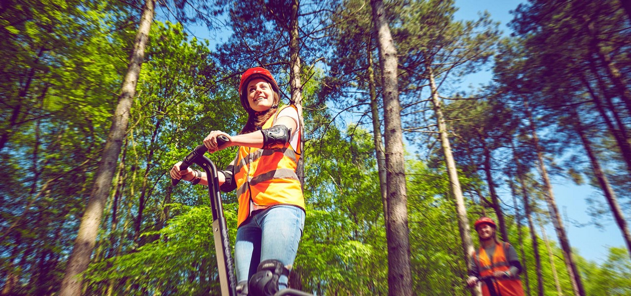 Two riders steer Segways, smiling, wearing orange vests, helmets, and pads, moving along a trail. Surrounding them, tall sunlit trees create a green forest canopy under a clear sky.