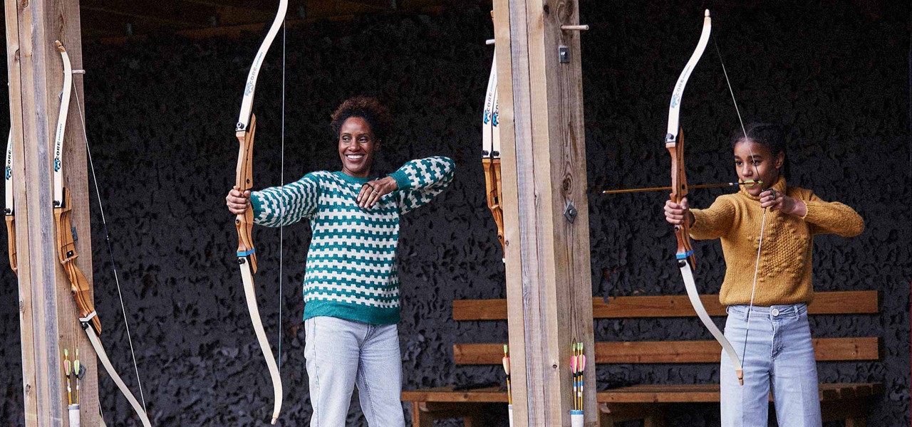 Archers shoot; one draws an arrow while another holds a bow smiling; covered wooden range with hanging bows, benches, and quivers. Text: CORE on multiple bows.