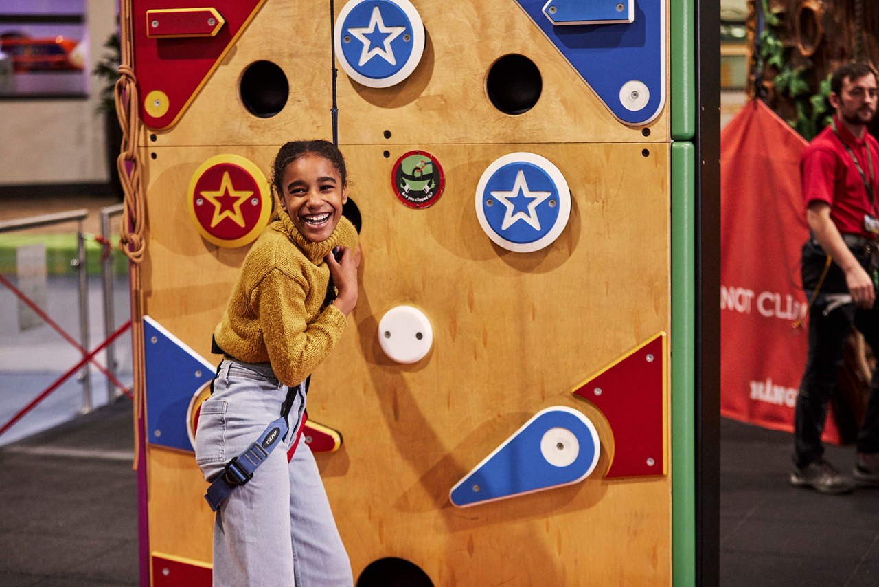 Child climber laughs, gripping a belay rope; colorful wall with star-decorated holds in an indoor climbing gym; an instructor stands near an orange safety banner.
Text: “NOT CLI...” (partial). Other small stickers have unreadable text.