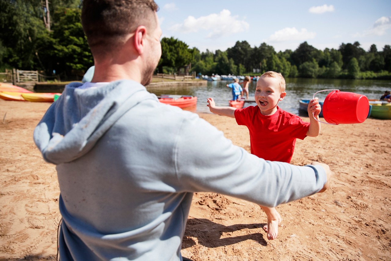 Child runs laughing toward an adult for a hug, swinging a red bucket; sandy lakeshore with kayaks, scattered people, calm water, and tree-lined background under a bright, blue sky.