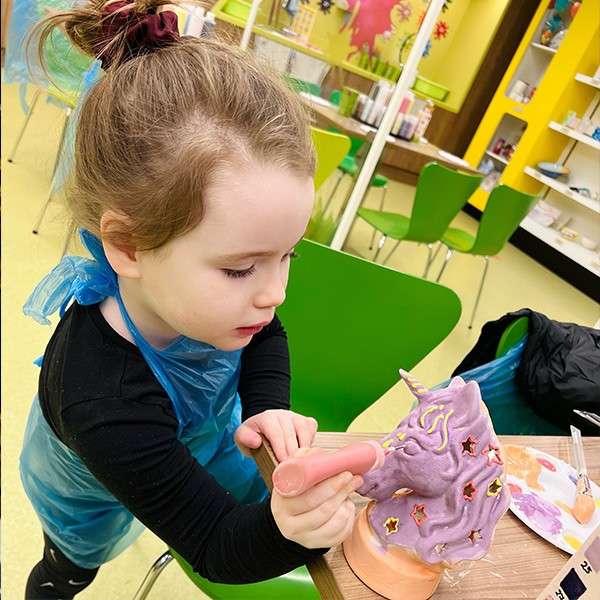 A little girl decorating a pottery unicorn head.